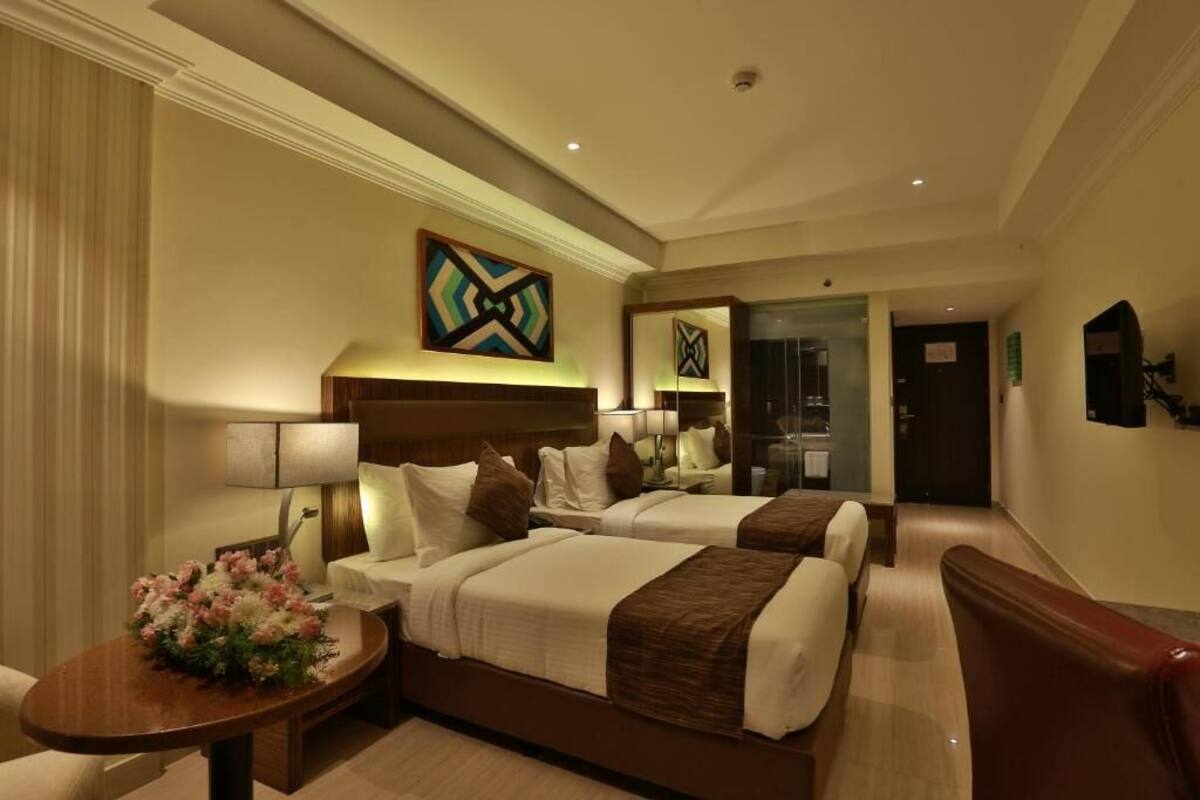 A hotel room with two beds, a desk with a lamp, a flower arrangement, and a painting on the wall.