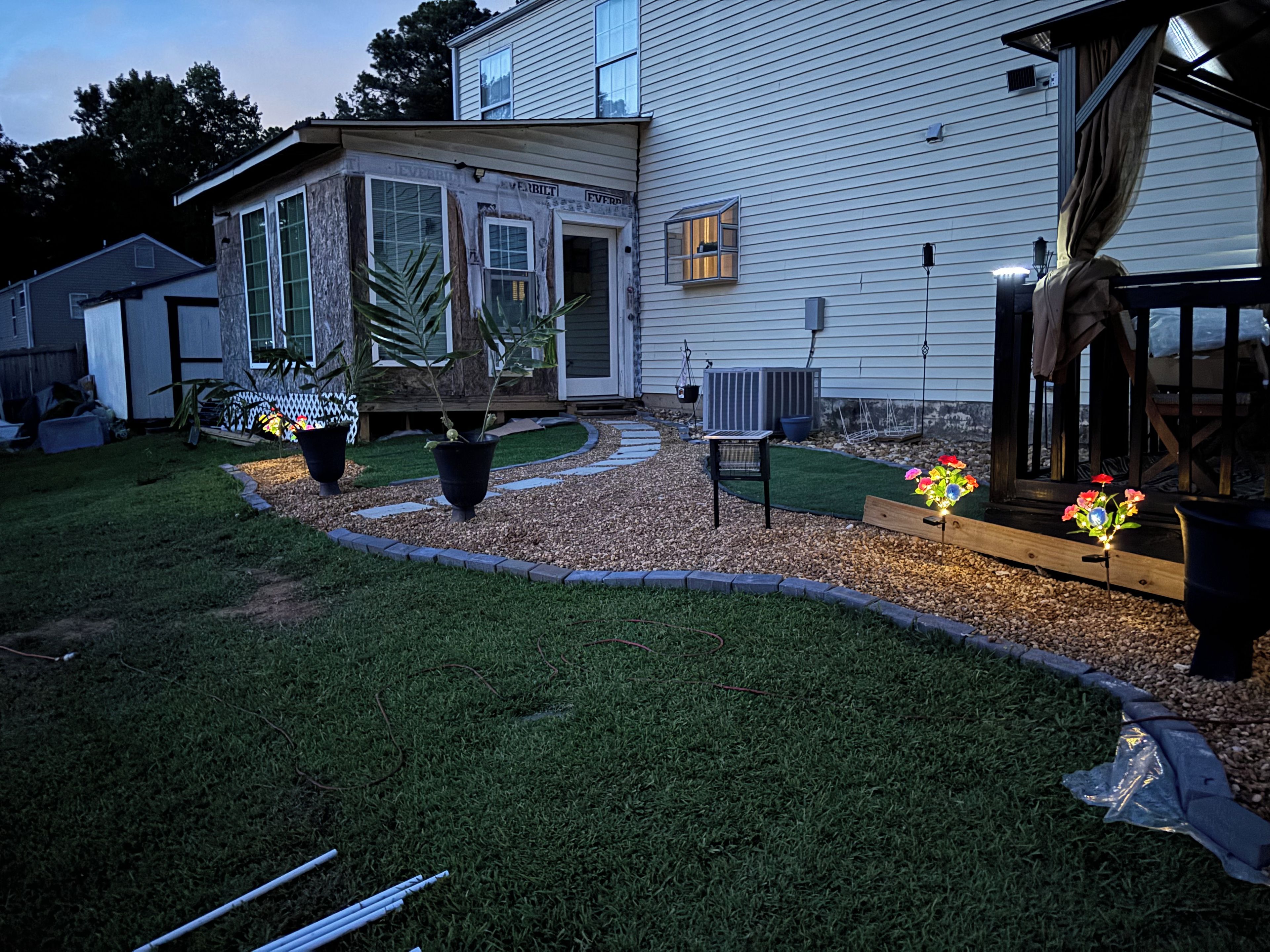 A residential property with a lawn, potted plants, a wooden deck, and a house with visible siding and windows.
