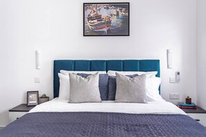 Quarto bem arrumado com cama, almofadas, quadro emoldurado, mesas de cabeceira e uma pequena planta.