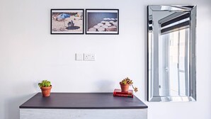 Ein modernes Zimmer mit einer dunklen Holzkombie, zwei Kübelpflanzen, zwei gerahmten Strandfotos und einem großen Spiegel, der einen hellen Raum widerspiegelt.
