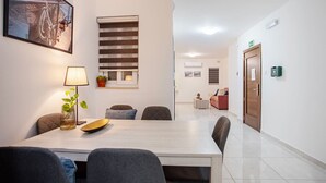 Een moderne hotelkamer met een zithoek, een groot raam met zonwering, een nachtkastje met lamp en een kleine plant.