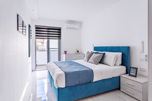 Une chambre moderne avec un lit bleu, des murs blancs et un balcon doté d’une porte coulissante.