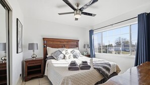 Ein Schlafzimmer mit einem Bett, Nachttischen, einem Deckenventilator, einem Fenster mit Vorhängen und Blick ins Freie.