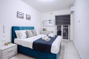 Quarto de hotel moderno com uma cama, mesa de cabeceira e uma cômoda pequena. Há uma janela com persianas, um ar condicionado fixado na parede e um quadro emoldurado.