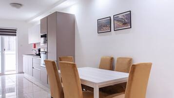 Un espace repas moderne avec une table blanche et quatre chaises, une cuisine avec des meubles blancs et deux tableaux encadrés au mur.