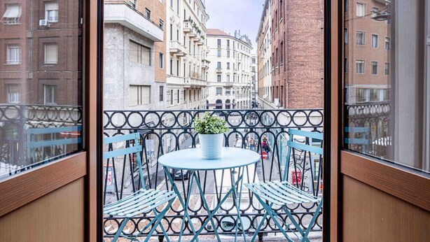 A balcony with a round table and chairs, a potted plant, and a view of a street with buildings. - Easylife - Milano - Mora 2 - Ticinese  (Milano)