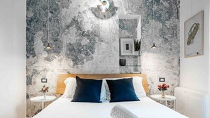 A modern hotel room with a large bed, two bedside tables, a nightstand, a wall-mounted mirror, and a textured wall. - Easylife - Milano - Mora 2 - Ticinese  (Milano)