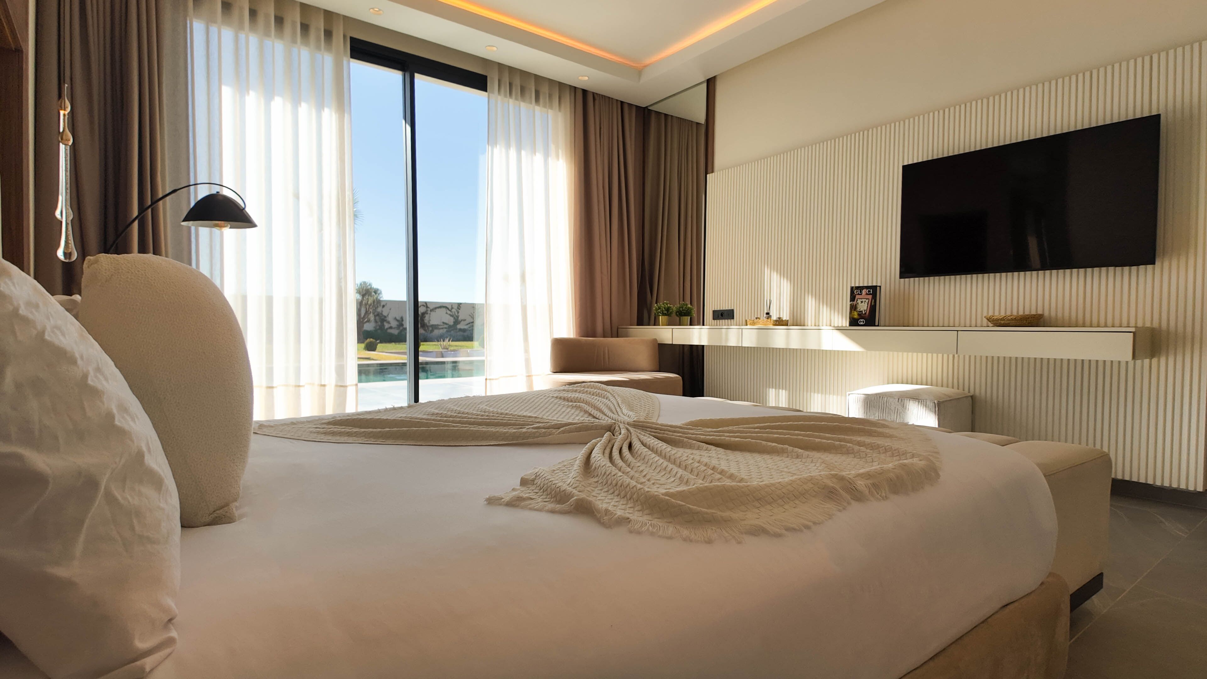 A modern hotel room with a large bed, a flat-screen TV mounted on the wall, and a view of a swimming pool through the window.