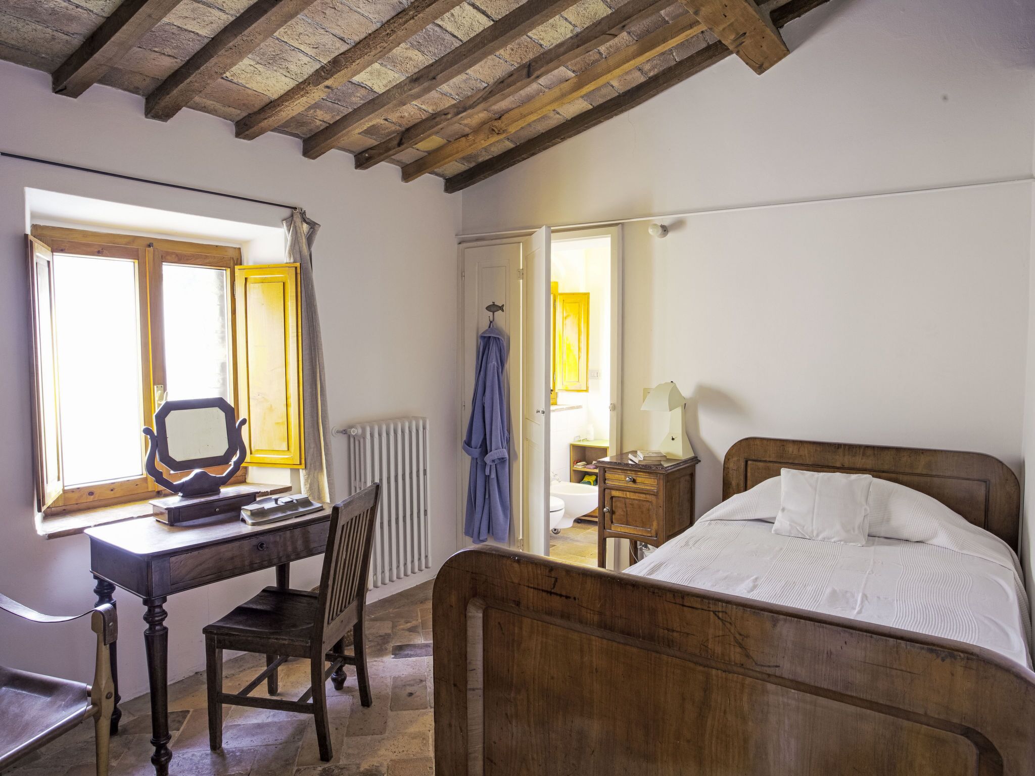 Une chambre à coucher avec un lit en bois, un bureau avec une chaise, une fenêtre et une salle de bain visible à travers une porte ouverte.