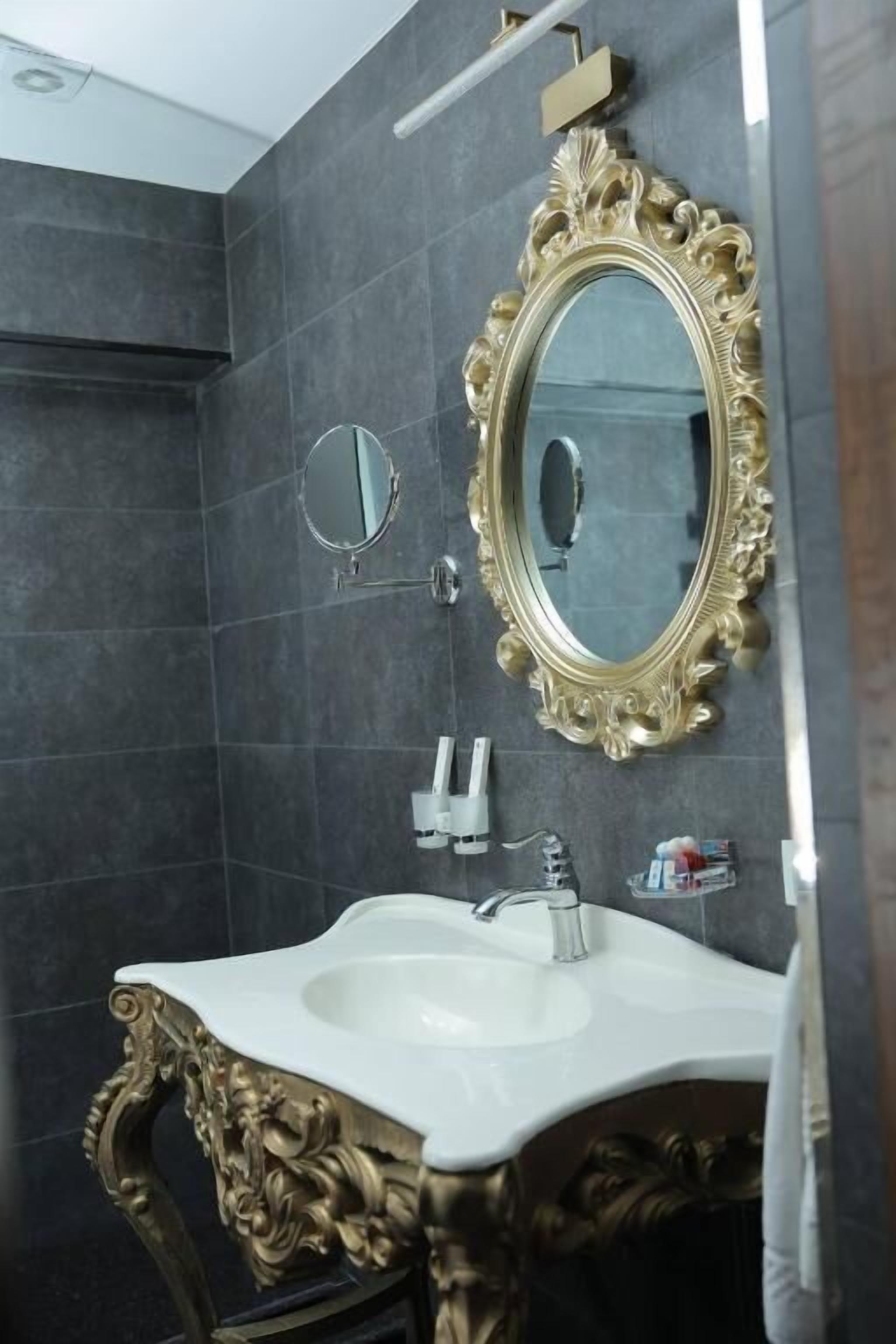 A bathroom with a large ornate mirror, a white sink, and a small round mirror on the wall.