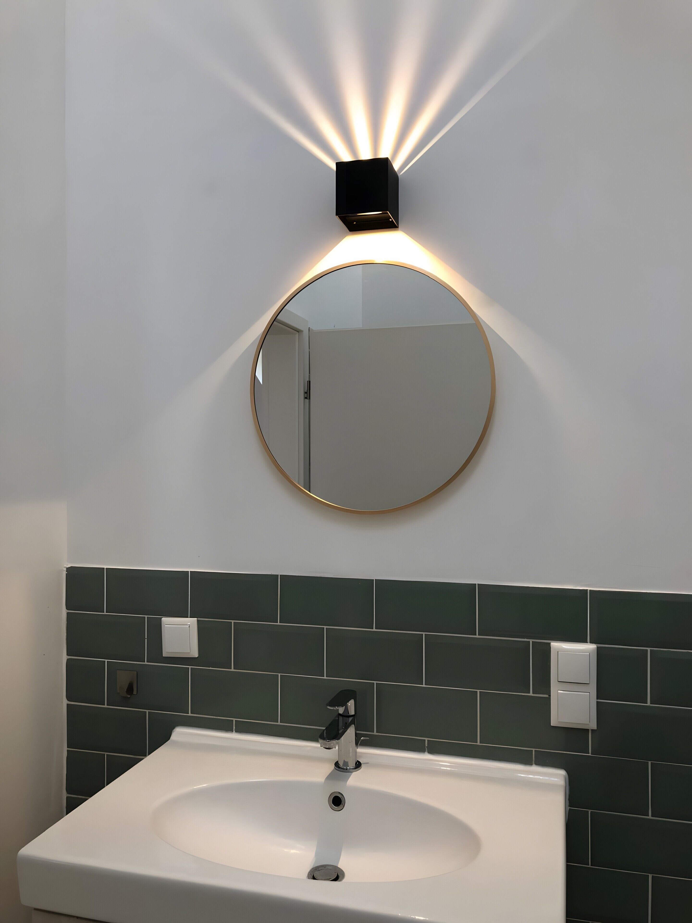 Ein Badezimmer mit einem weißen Waschbecken, einem runden Spiegel mit goldener Rahmung und einer wandmontierten Leuchte.
