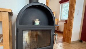 Moderni puukamiina, jossa on lasiluukku, on sijoitettu huoneeseen, jossa on puulattia ja ikkunasta avautuu näkymä punaiseen verhoon.