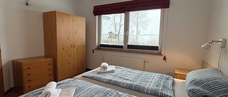 A bedroom with two single beds, a wooden wardrobe, a radiator, a window with a view, and a hanging light fixture.