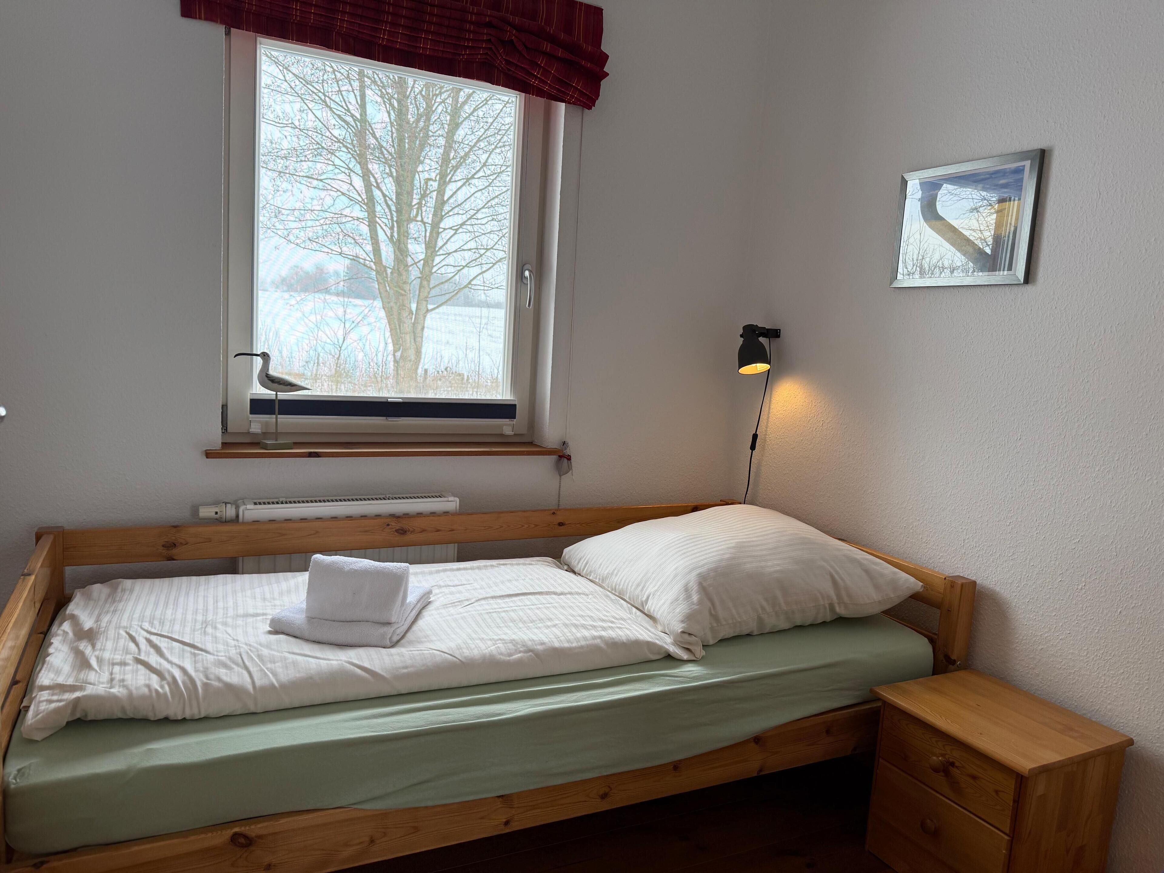 Een eenpersoonsbed met houten frame, een nachtkastje, een wandlamp, een raam met uitzicht op bomen en een ingelijst kunstwerk aan de muur.