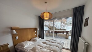 A hotel room with a wooden headboard, a bed with white bedding, a hanging lamp, a window with a view of a snowy landscape, and a balcony with a railing. - Apartment "Les Loges Blanches 101" with Wi-Fi (Châtel)