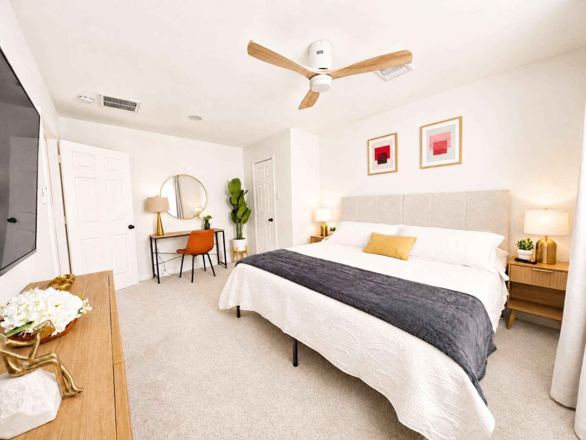 A hotel room with a bed, bedside tables, a desk, a chair, a ceiling fan, and artwork on the walls.