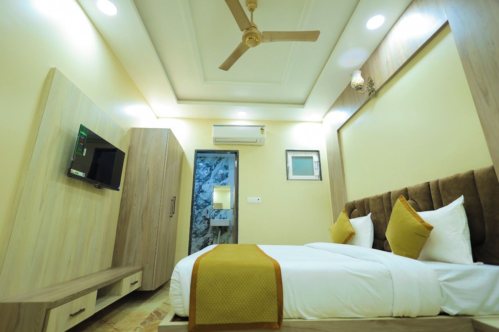 A hotel room with a bed, wardrobe, television, air conditioning unit, and a ceiling fan.