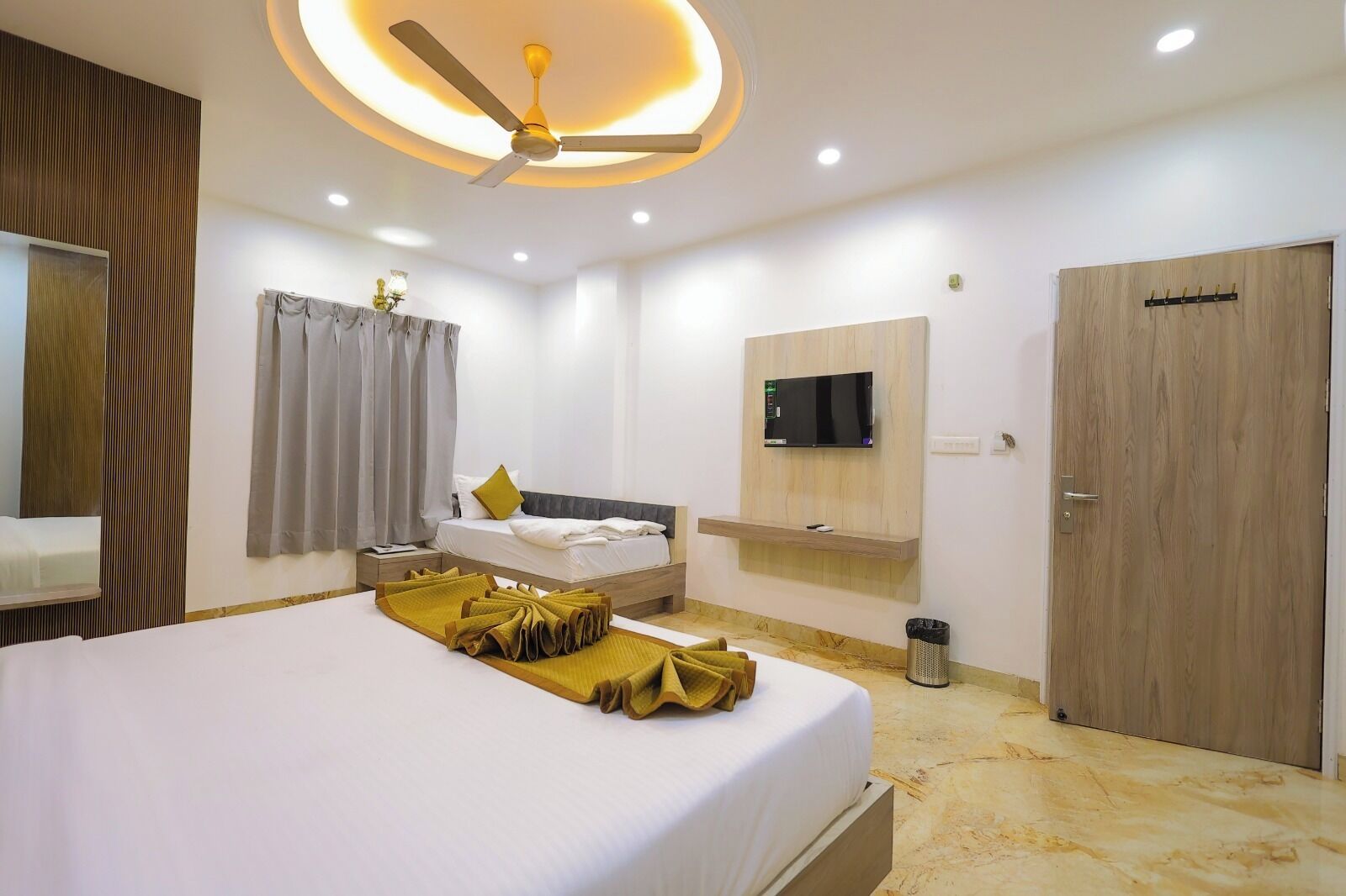 A modern hotel room with a large bed, a ceiling fan, a mounted TV, and a wooden door.