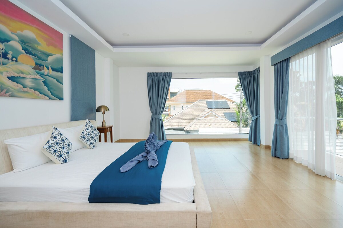 Ein modernes Hotelzimmer mit einem großen Bett, einem Gemälde an der Wand und Blick auf Gebäude durch das Fenster.