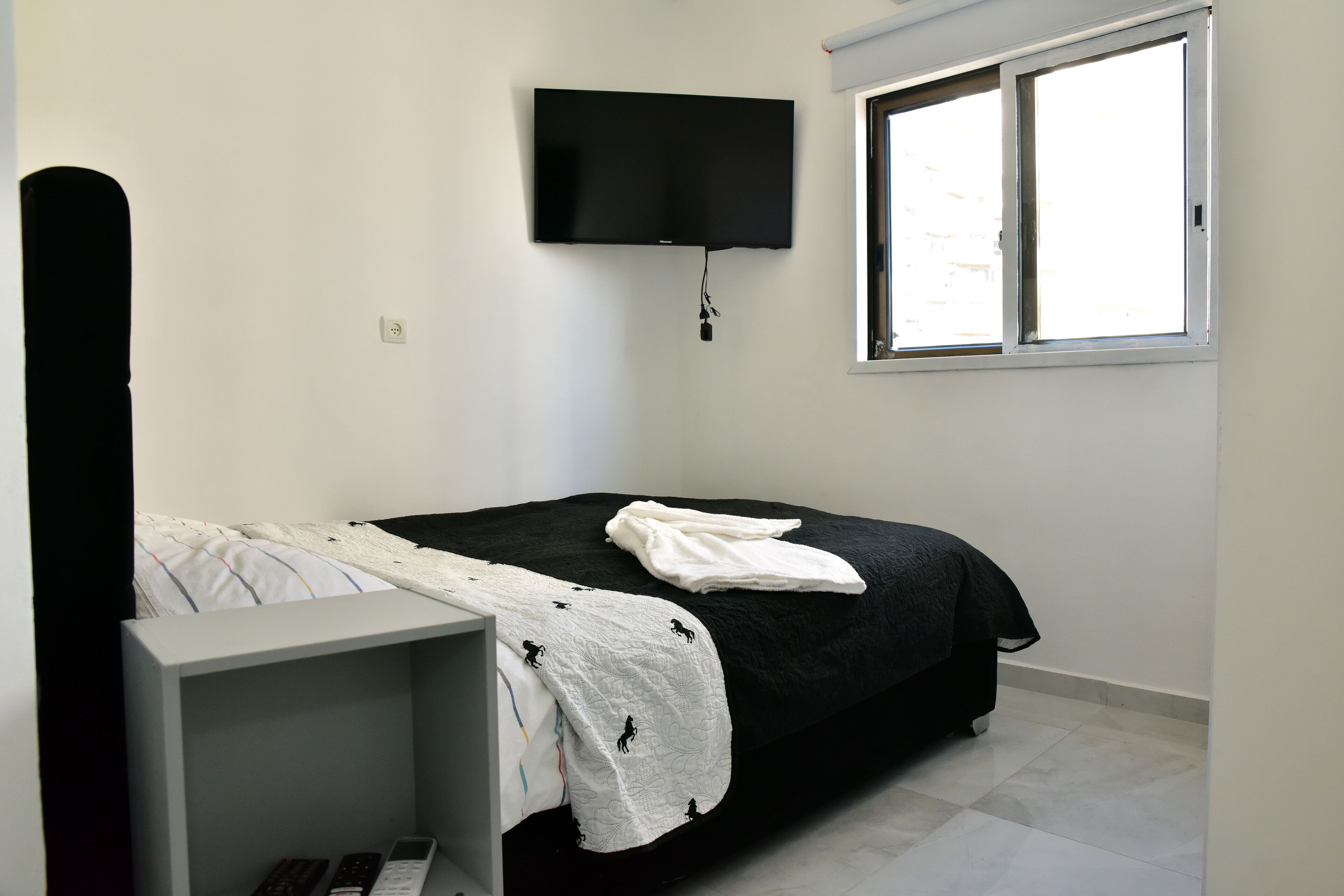 Une chambre à coucher avec un lit, un téléviseur fixé au mur, une fenêtre et une table de chevet.