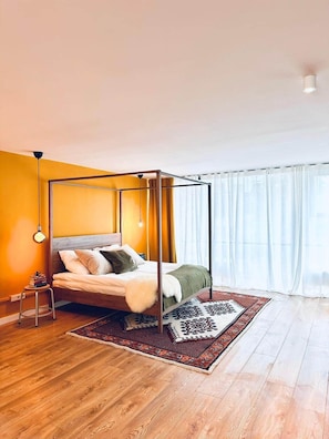 Un dormitori modern amb un llit amb cortines, un sòl de fusta, una catifa amb dibuixos i una finestra amb cortines primes.