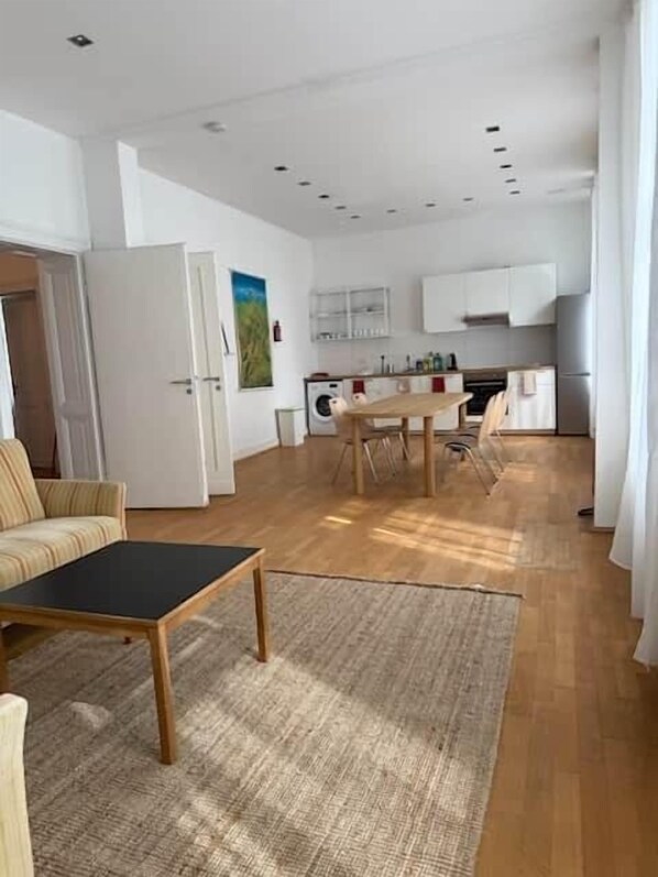 A modern living room with a sofa, a coffee table, and a rug. There is a dining area with a table and chairs, and a kitchen with cabinets and appliances.