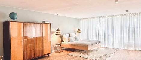 A bedroom with a wooden wardrobe, a bed with a white mattress, and a large window with sheer curtains.