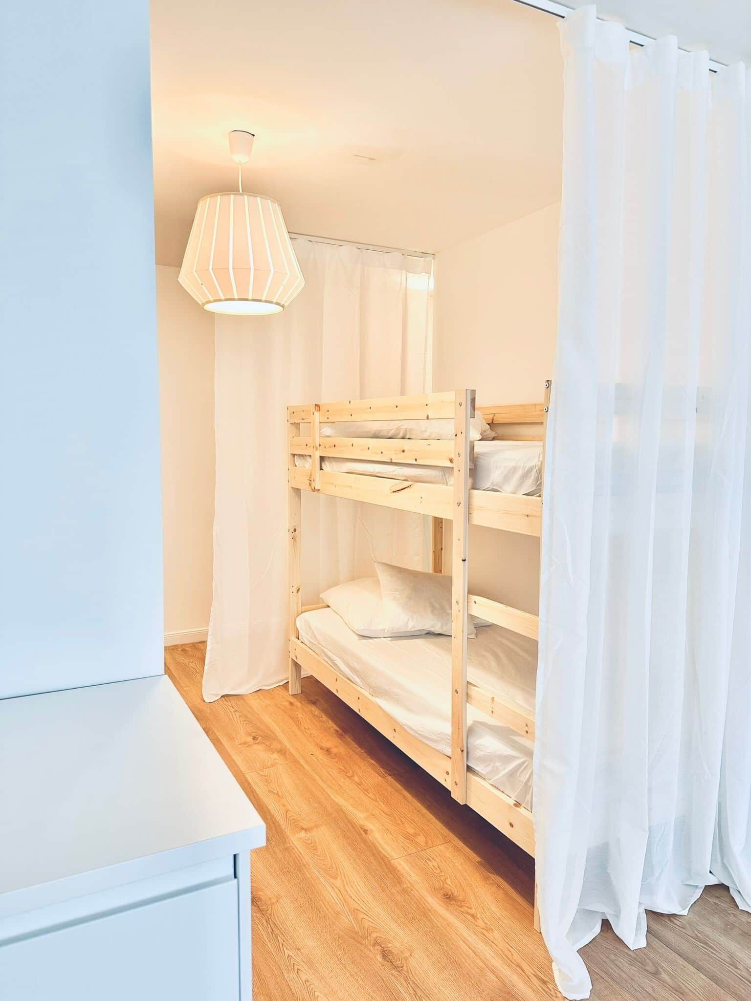 Dormitorio con literas, suelo de madera, paredes blancas y una lámpara colgante.