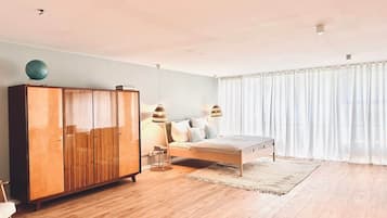 Ein Schlafzimmer mit einem Holzkasten, einem Bett mit weißer Matratze und einem großen Fenster mit leichtem Vorhang.