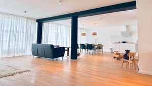 A spacious living area with a dark sofa, a dining table with chairs, and a kitchen area with a teddy bear on a chair. - Emmas Lofts - City center location (Essen)