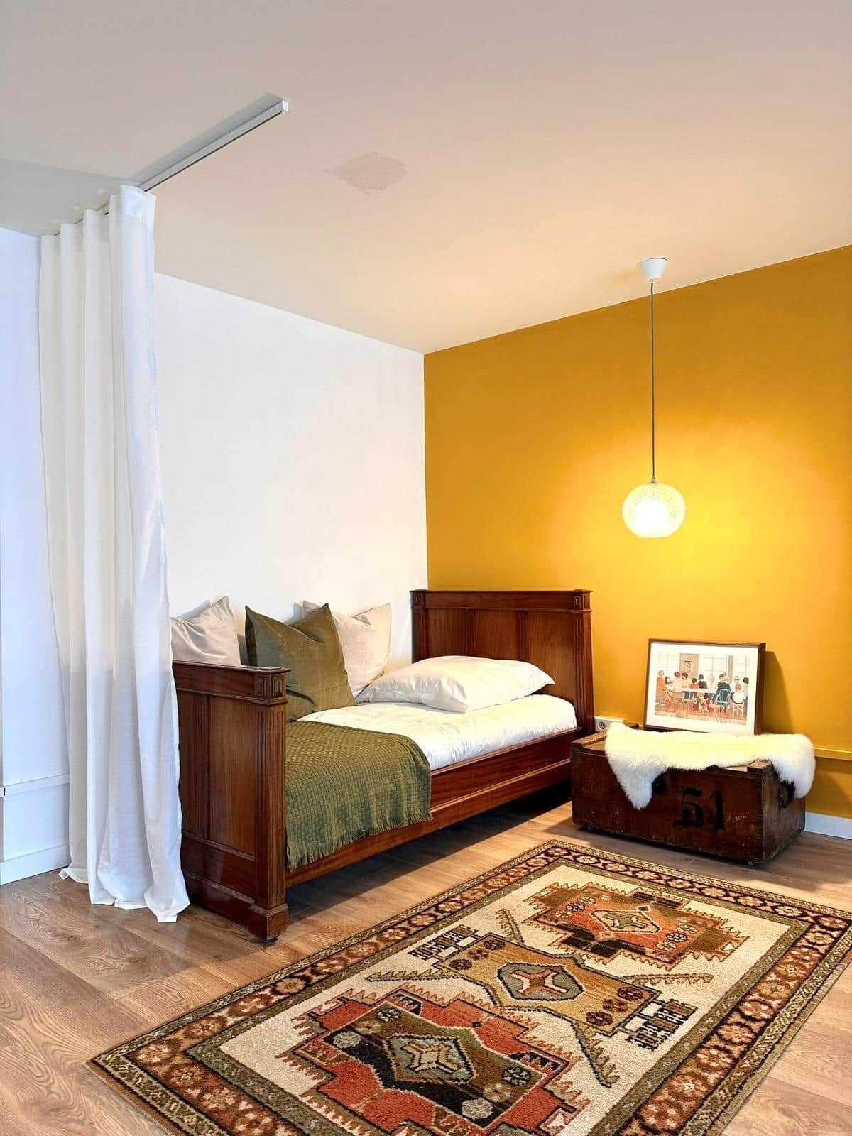 Ein Schlafzimmer mit einem hölzernen Bett, weißer Bettwäsche, einer gelben Akzentwand, einer Pendelleuchte, einem gemusterten Teppich und einem Vintage-Koffer.