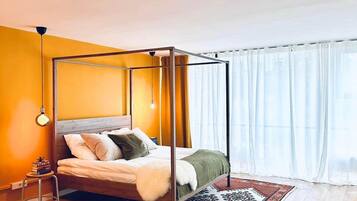 A modern bedroom with a four-poster bed, a wooden floor, a patterned rug, and a window with sheer curtains.