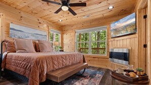 Camera da letto in una baita di legno, con un letto, una ventola a soffitto, una TV e una finestra con vista sugli alberi.