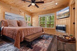 Ein Schlafzimmer einer Holzhütte mit Bett, Deckenventilator, Fernseher und Fenster mit Blick auf Bäume.