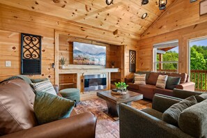 A cozy cabin interior with a fireplace, comfortable seating, and a large window overlooking greenery. - Majestic Horizon by Ghosal Luxury Lodging (Sevierville)