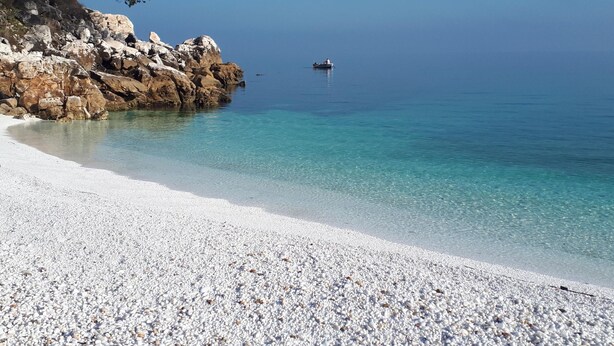 Ein Kiesstrand mit klarem Wasser, eine felsige Klippe und ein Boot in der Ferne.