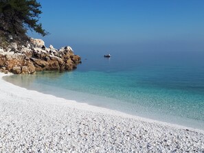 Ein Kiesstrand mit klarem Wasser, eine felsige Klippe und ein Boot in der Ferne.