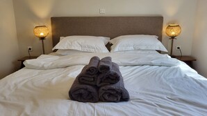 Ein ordentlich bezogenes Bett mit weißen Leinentüchern, zwei Nachttischlampen und ein Stapel zusammengerollter Handtücher in der Mitte.