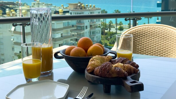 Een tafel met ontbijtartikelen, zoals sinaasappels, croissants en sapglazen, met uitzicht op de kust.