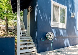 Une maison bleue avec une porte blanche, une fenêtre avec des stores, un porche avec une balustrade blanche et un tuyau d’arrosage raccordé à la maison.