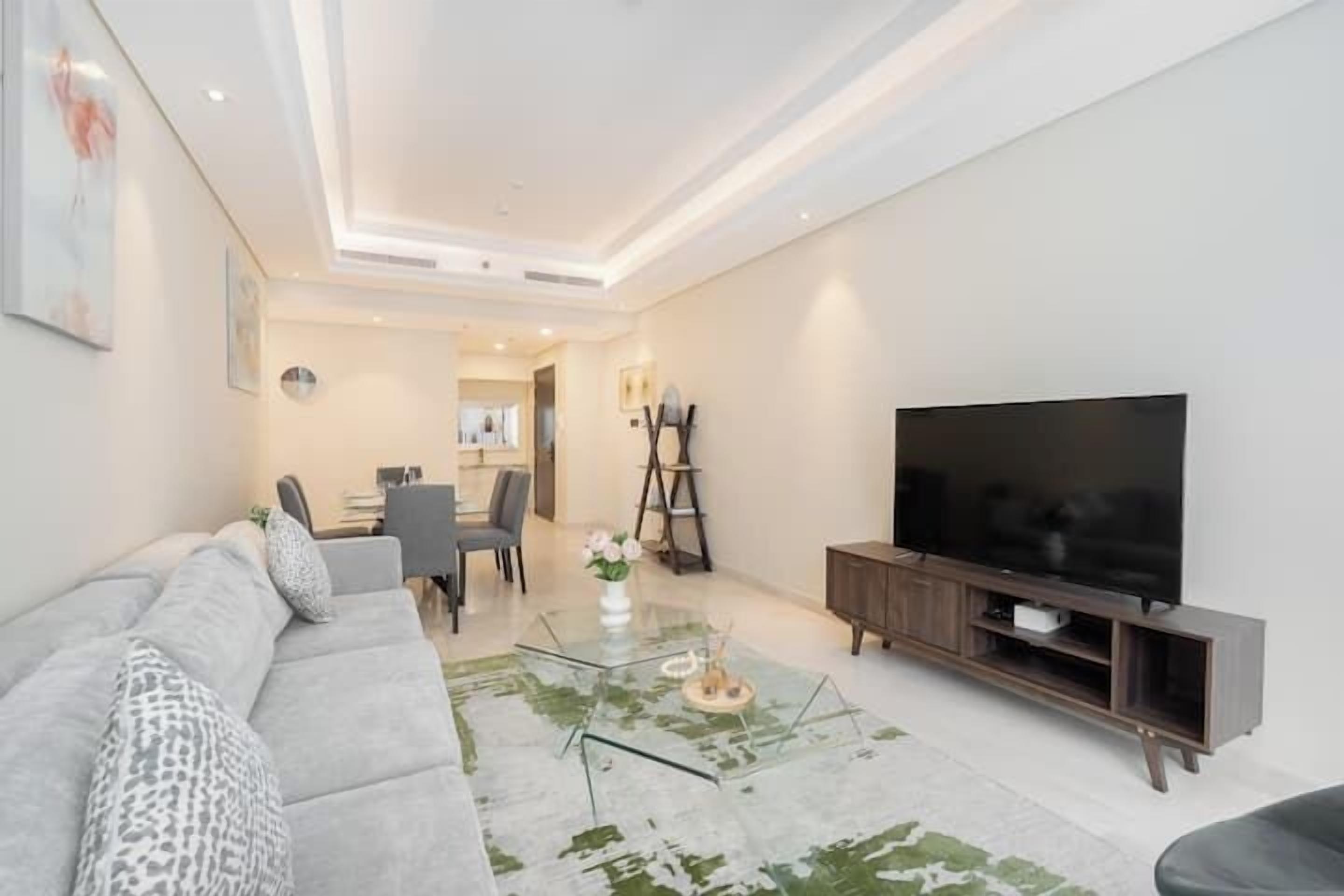 A modern living room with a grey sofa, a glass coffee table, a flat-screen TV, and a dining area in the background.