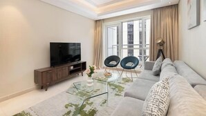 A modern living room with a sofa, two chairs, a glass coffee table, a TV on a wooden cabinet, and a large window with curtains. - Vogue | Mon Reve Downtown near Dubai Mall | Burj Khalifa (Dubai)