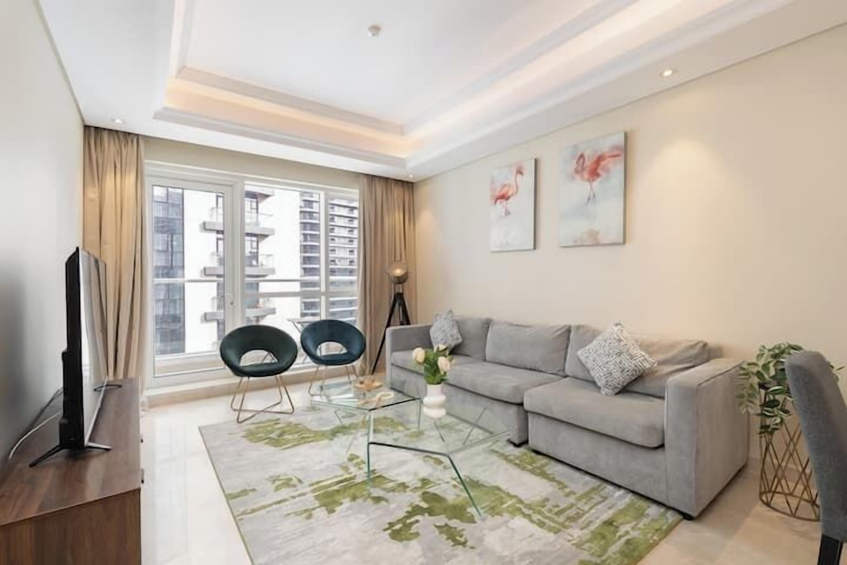 A modern living room with a grey sofa, two black chairs, a glass coffee table, and a large window with a view of high-rise buildings.