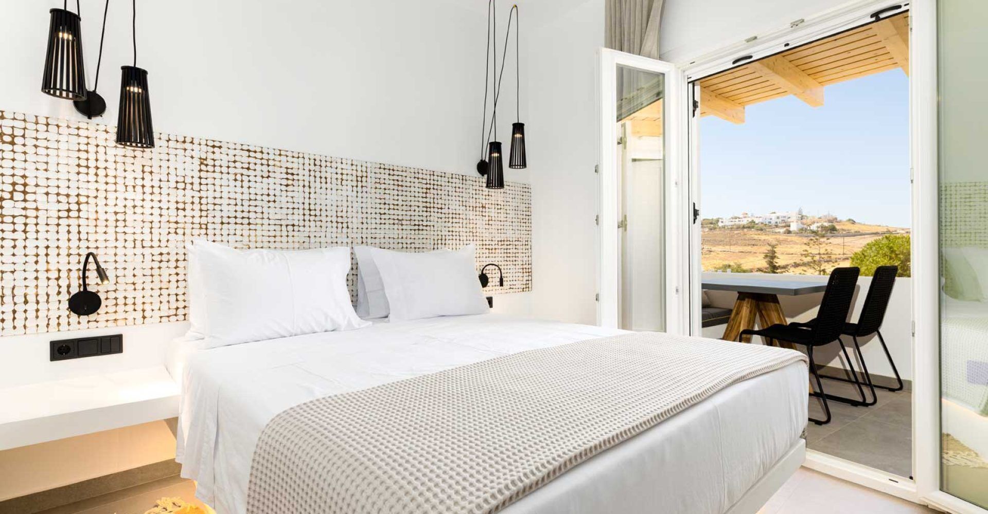 A modern bedroom with a large bed, a headboard with a woven pattern, and a view of a landscape through an open door.