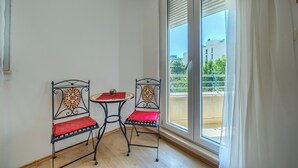 Una habitación con dos sillas de metal y una mesita pequeña, una puerta corrediza de vidrio a un balcón exterior, y vistas de edificios y vegetación.