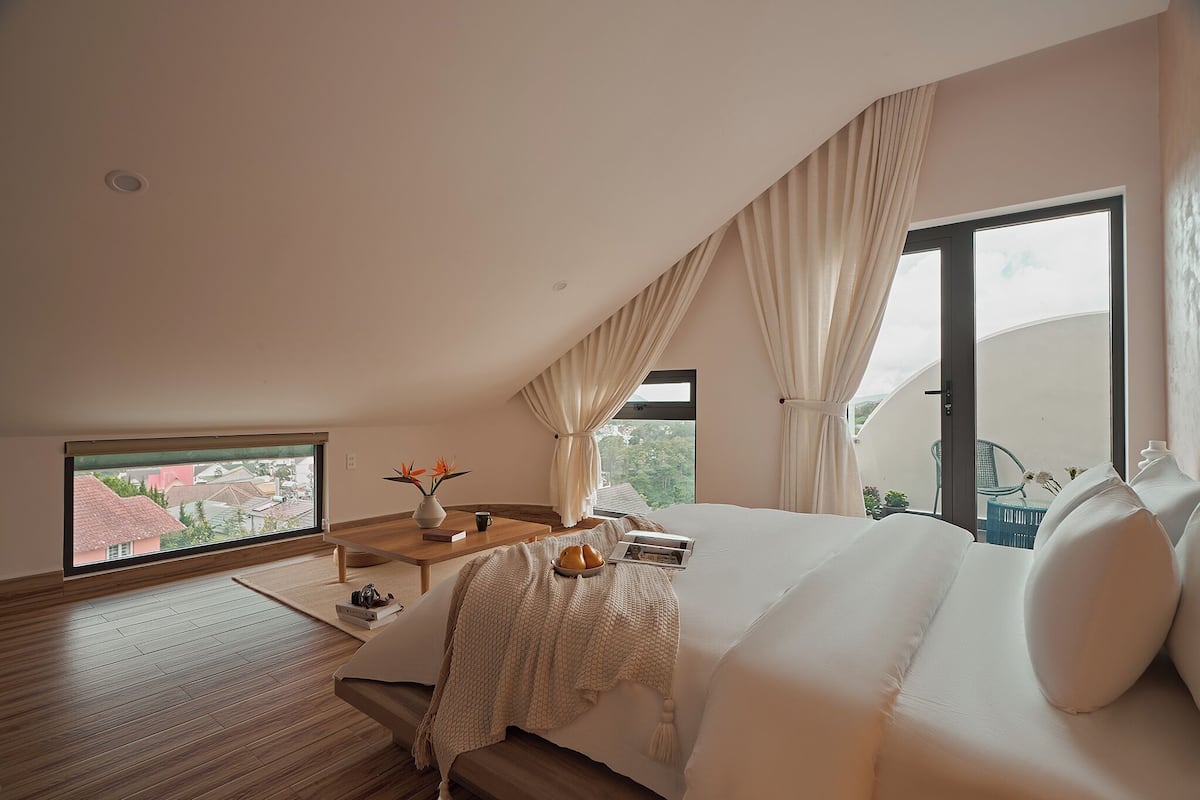 Une chambre avec un grand lit, une fenêtre avec des rideaux, une petite table sur laquelle est posé un vase, et une vue sur l’extérieur.