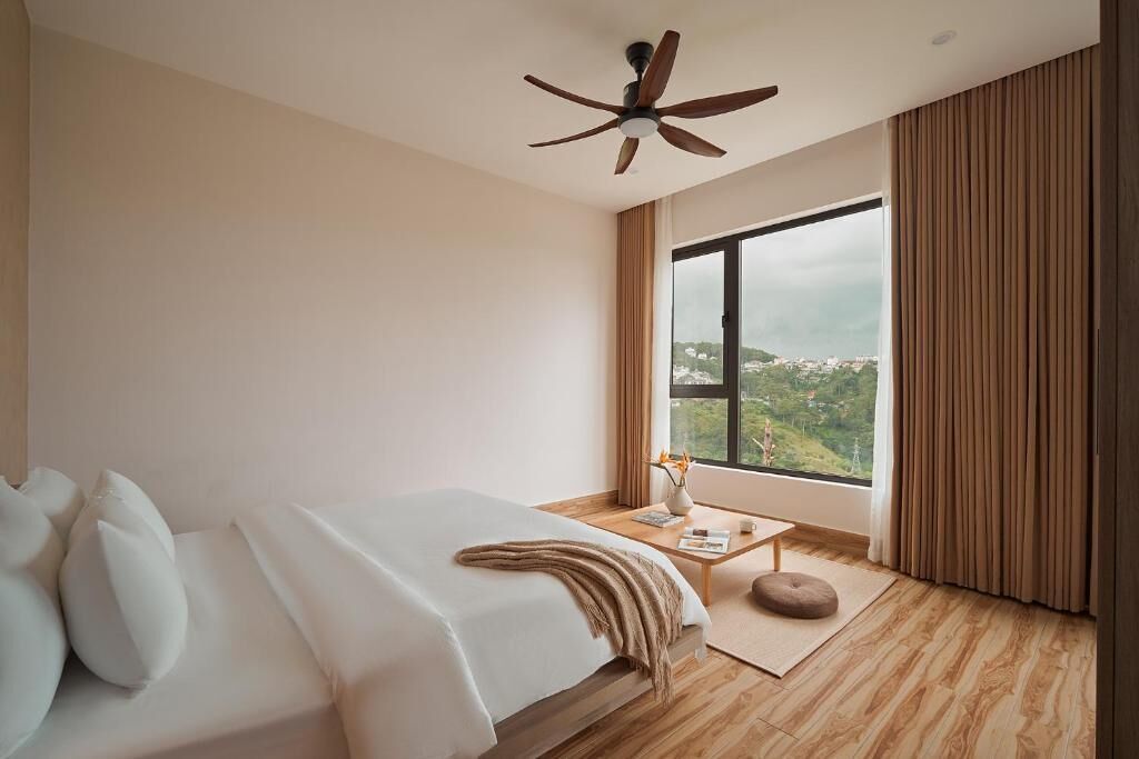 A modern hotel room with a large bed, a ceiling fan, a view of a cityscape, and a window with curtains.