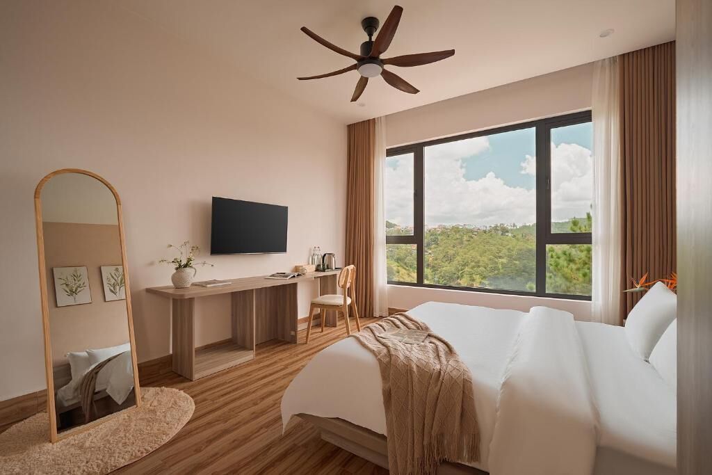 A modern hotel room with a large bed, a desk with a flat-screen TV, a ceiling fan, and a large window offering a view of the outdoors.