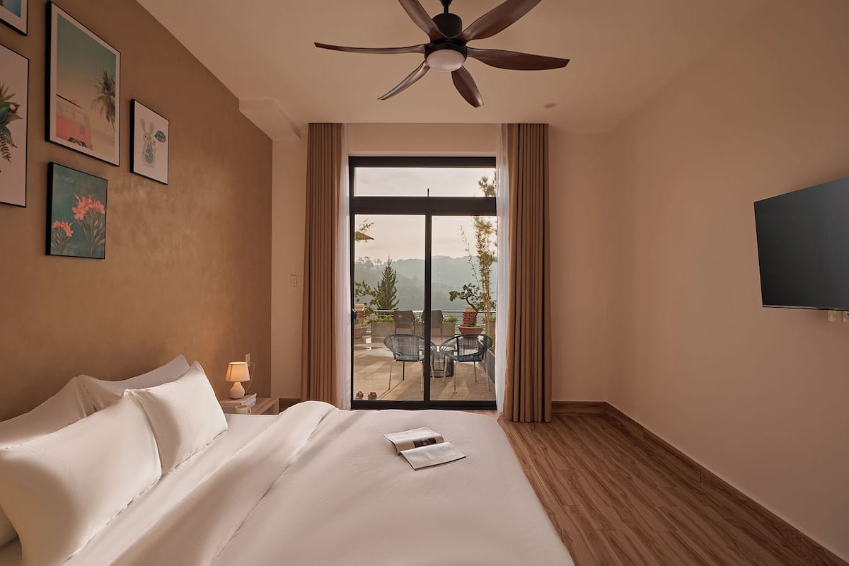 Une chambre d’hôtel avec un lit, un ventilateur de plafond, une télévision et un balcon équipé de chaises et d’une table.