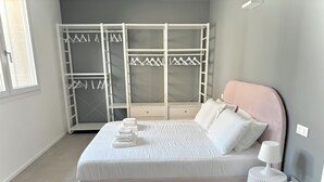 Een slaapkamer met een bed, nachtkastje, lamp en een kledingkast met daarin hangende kleding.